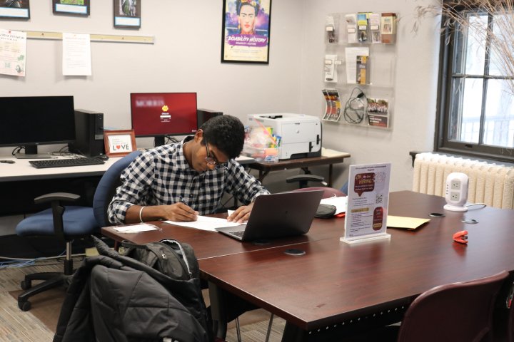 A student sits at a desk in a computer lab, focused on writing with papers spread out in front of them. A laptop is open on the desk, and the room has posters, a printer, and office supplies in the background.